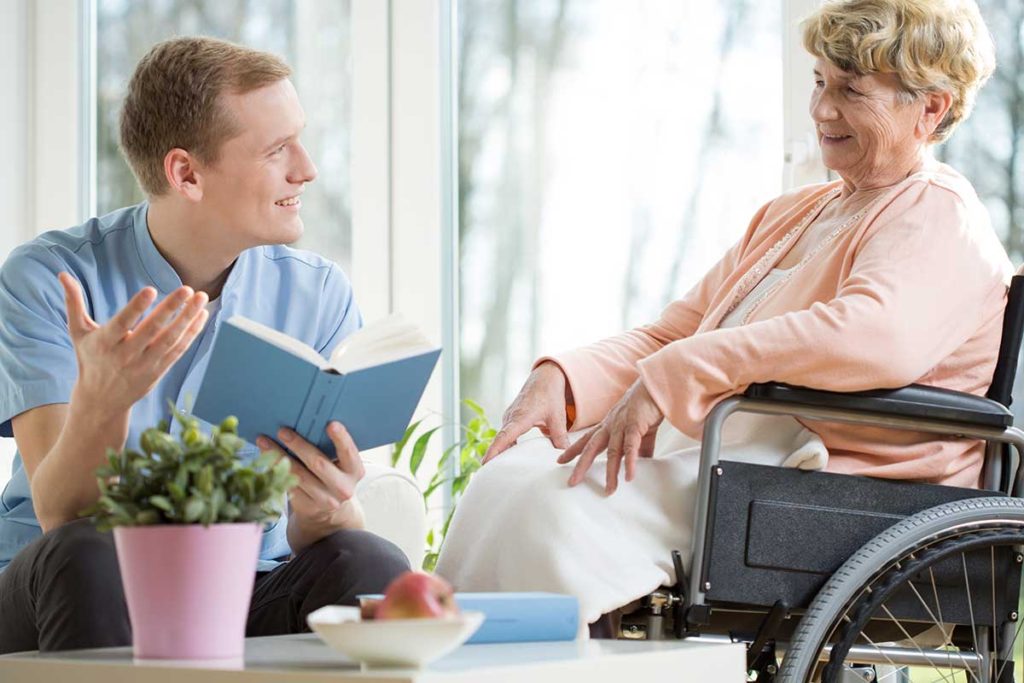 Caregiver reads to a smiling elderly woman in a wheelchair by a sunlit window.