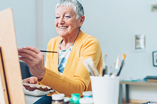 Elderly woman painting joyfully in a bright room with art supplies around her.