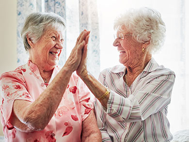 Two elderly women smiling and high-fiving in a sunlit room.