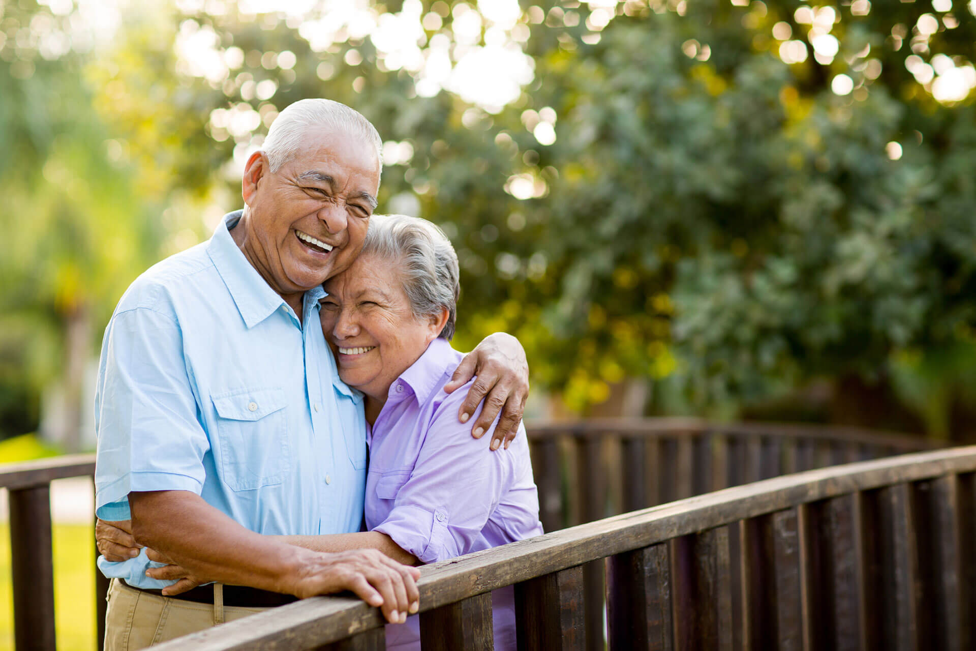 Smiling elderly couple embracing on a wooden bridge in a lush garden.