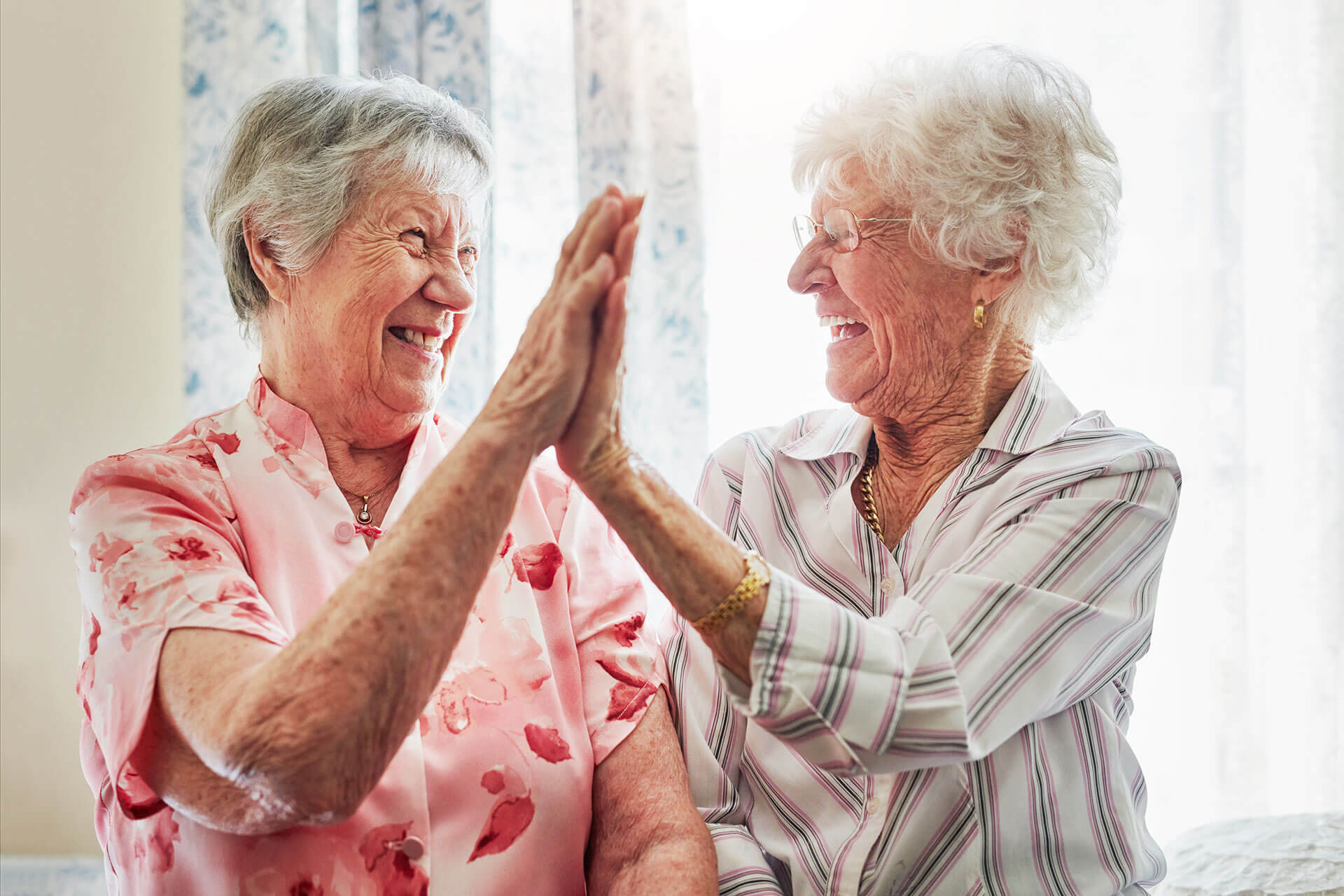 Two elderly women smiling and giving each other a high-five indoors.