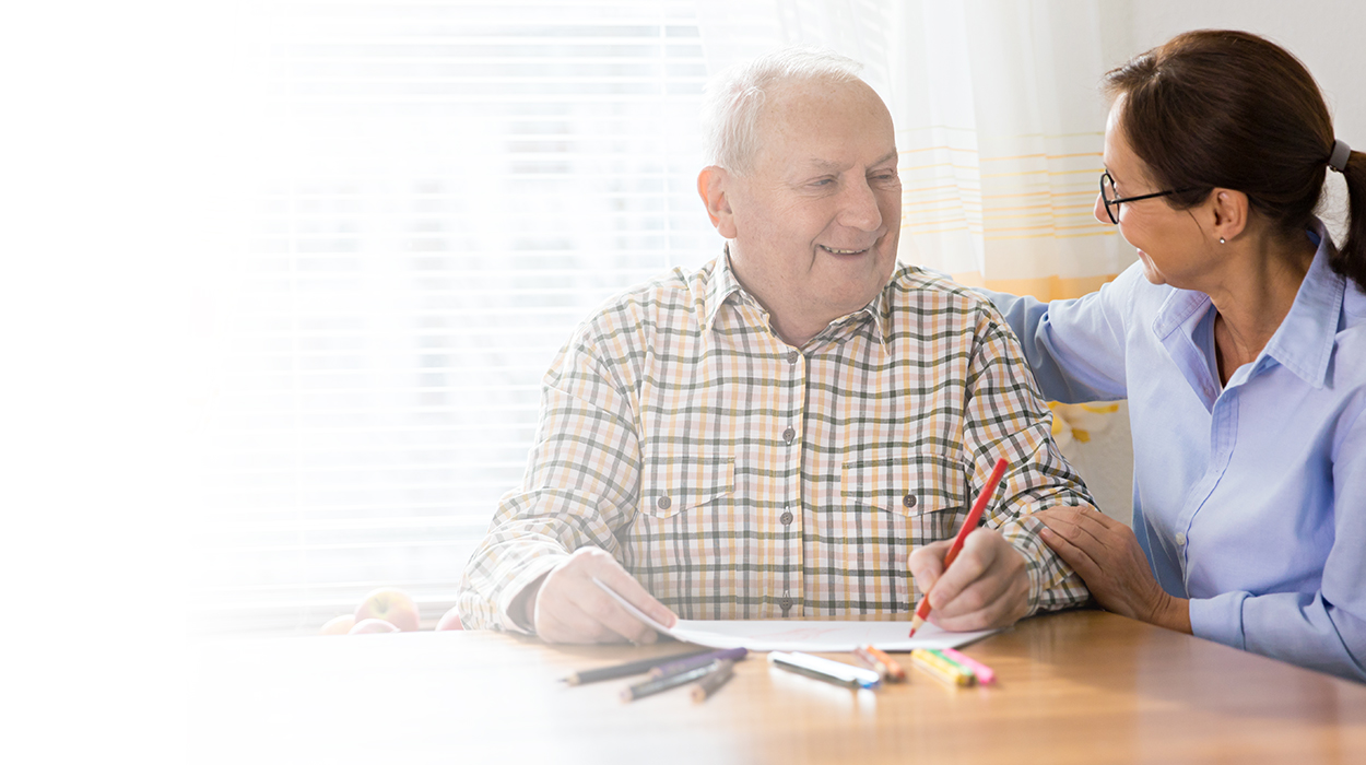 Elderly man in plaid shirt smiling with supportive caregiver at table.