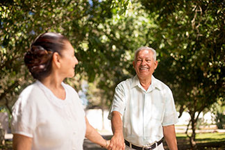 Smiling couple walking hand in hand through a sunlit garden area with trees.