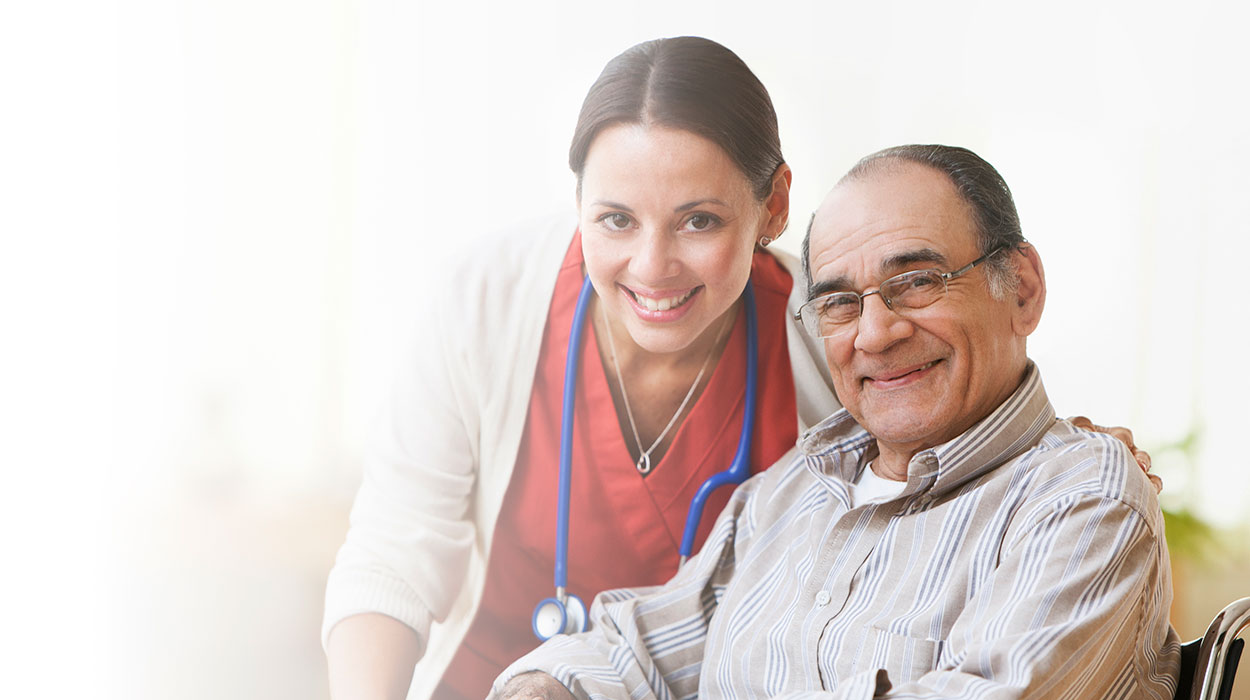 Nurse with stethoscope smiling beside seated elderly man indoors.