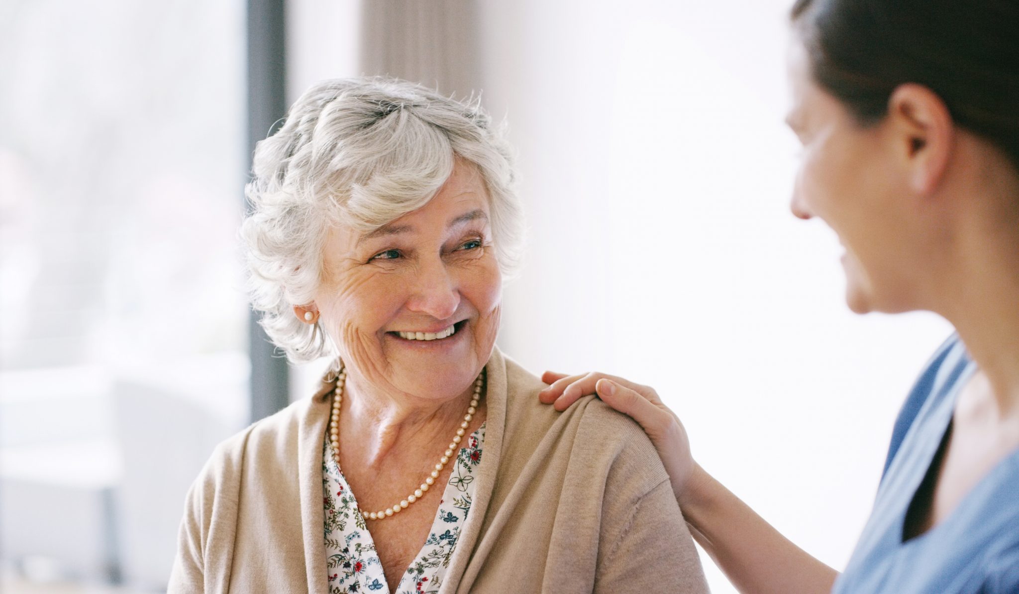 Elderly woman smiling at caregiver with hand on her shoulder.