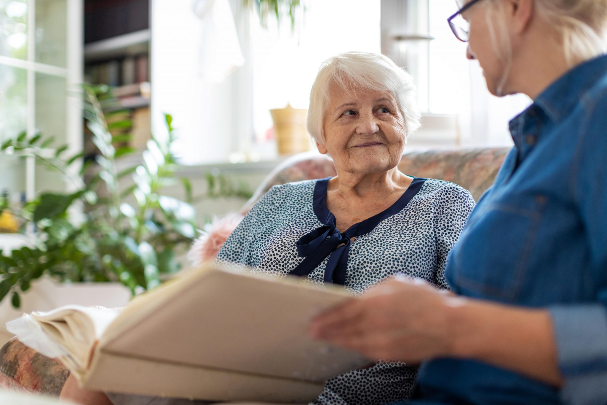 Elderly woman talking with a caregiver on a cozy couch in a bright living room.