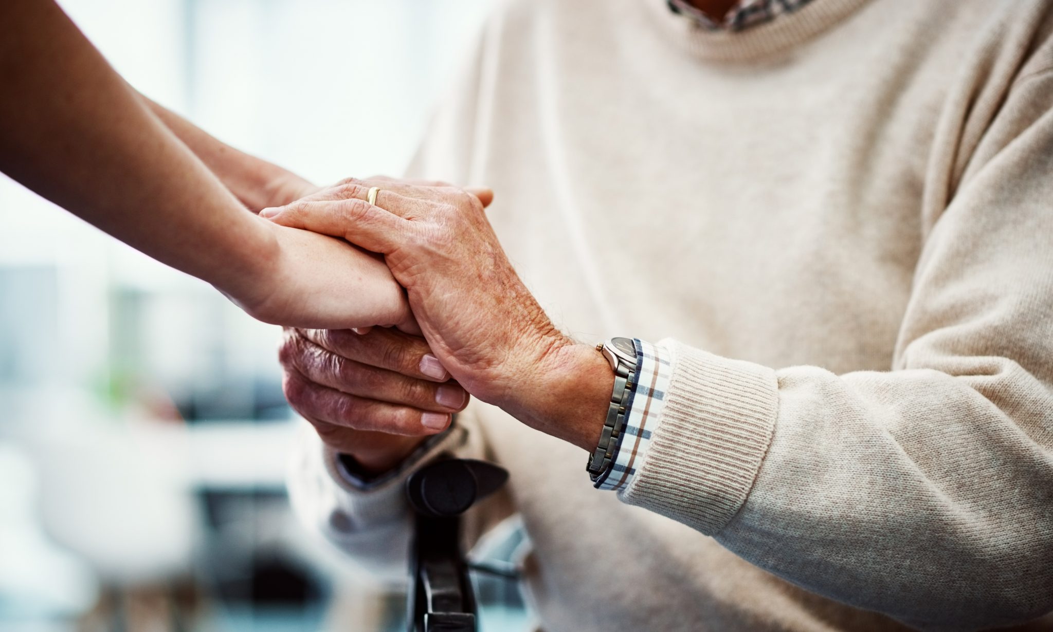 Elderly person in a sweater holding hands with a younger individual in a comforting gesture.