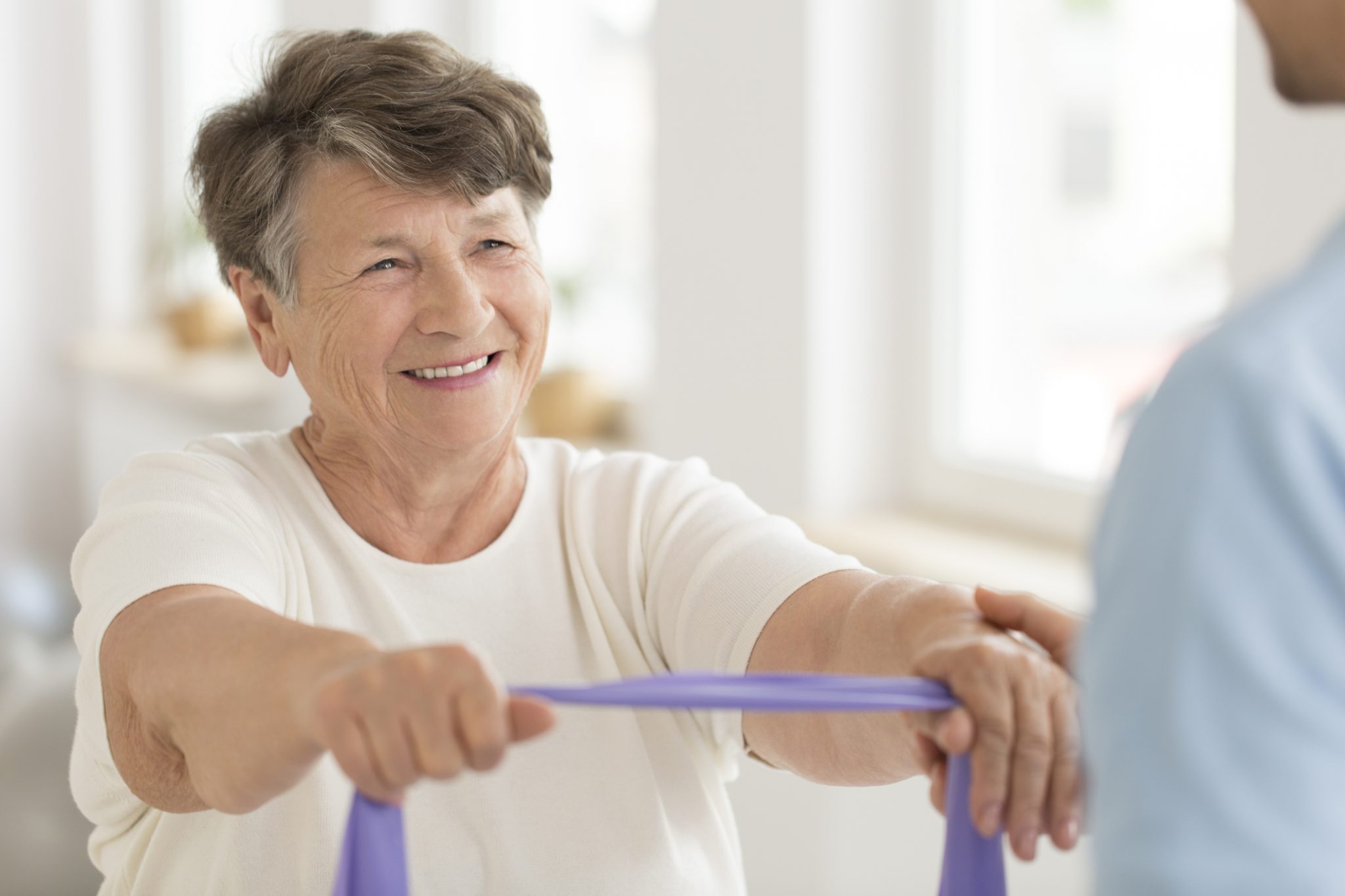 Elderly woman smiling and holding a purple resistance band during a seated exercise.