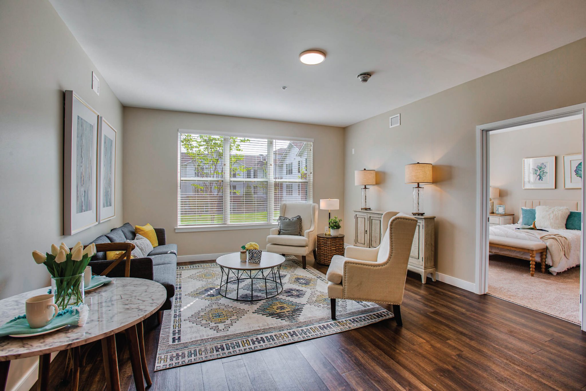 Cozy living room with modern furniture, large window, and adjacent bedroom visible through open door.